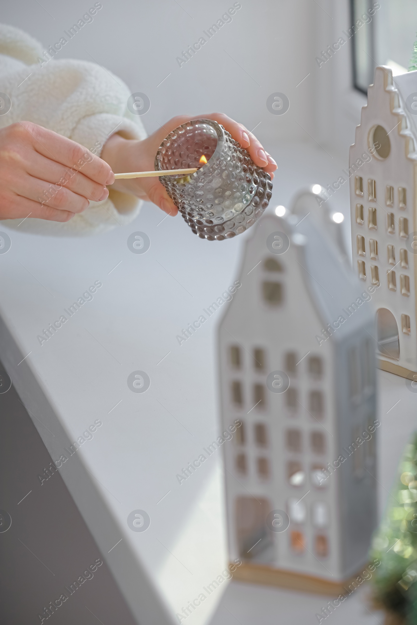 Photo of Woman lightning up candle near windowsill with house shaped lanterns indoors, closeup