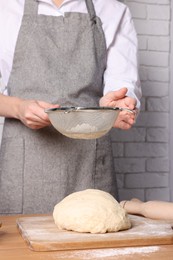 Woman sprinkling flour over dough at table near white brick wall, closeup