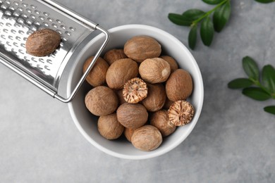 Nutmegs in bowl, metal grater and green branches on light grey table, flat lay