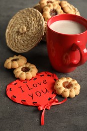 Photo of Heart shaped greeting card with phrase I Love You near cookies and red cup on dark grey table