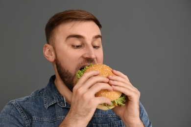 Young man eating tasty burger on grey background. Space for text