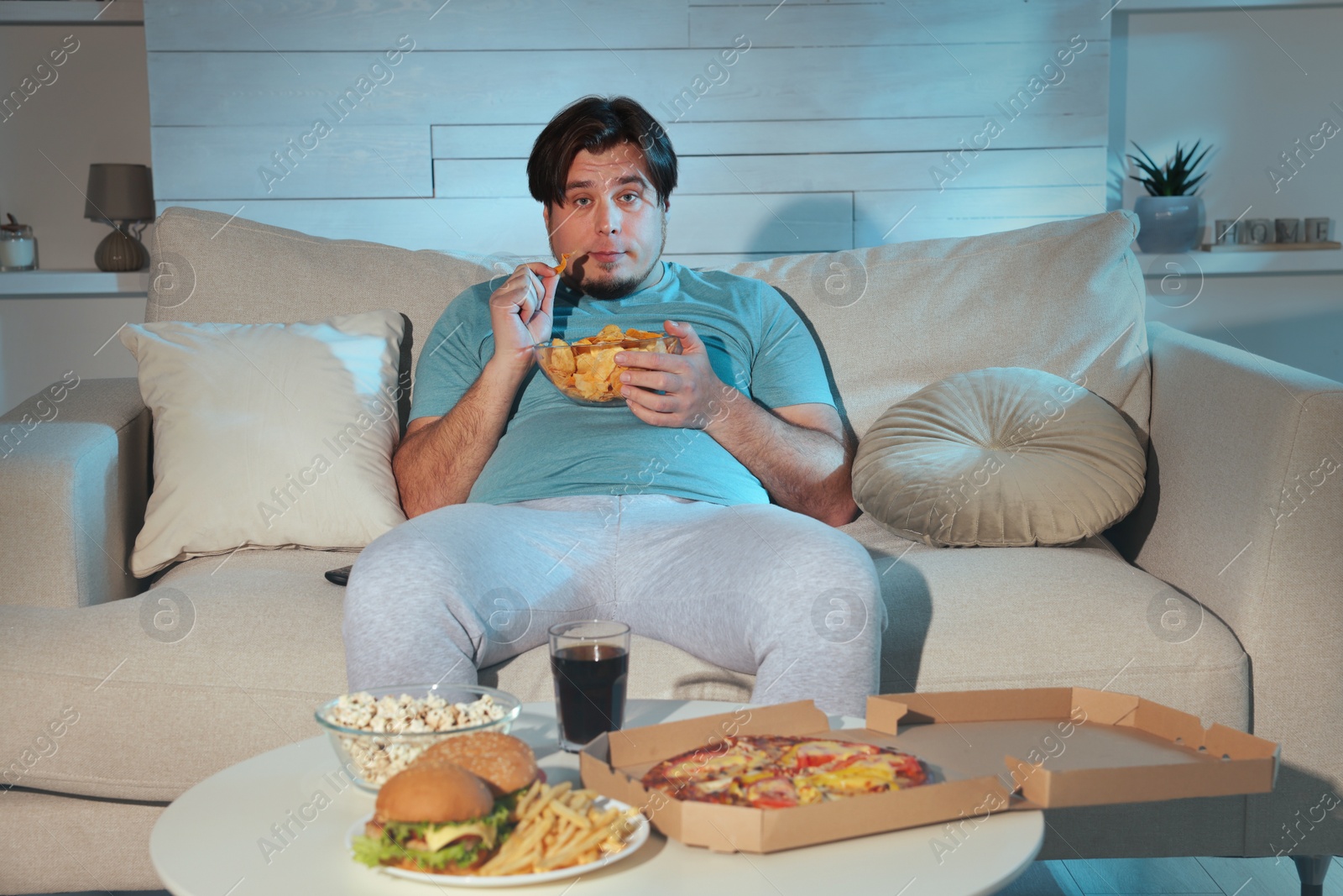 Photo of Overweight man with chips watching TV on sofa at home