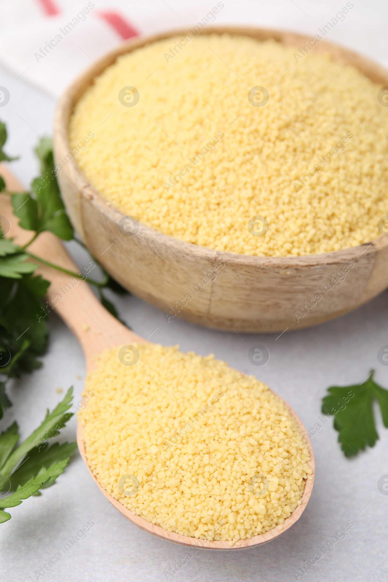 Photo of Raw couscous in bowl, spoon and parsley on light table, closeup