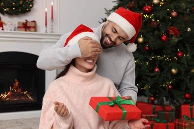 Happy man in Santa hat surprising his girlfriend with Christmas gift at home