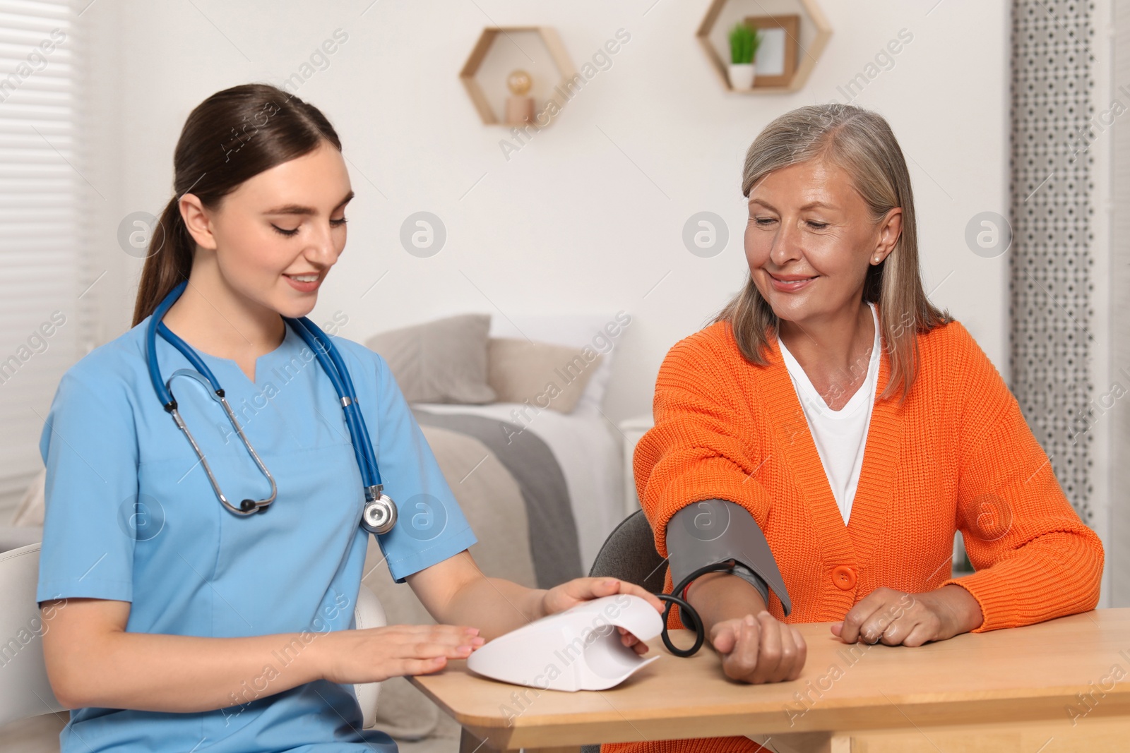 Photo of Young healthcare worker measuring senior woman's blood pressure at wooden table indoors