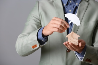 Photo of Male agent covering wooden house with umbrella cutout on grey background, closeup. Home insurance