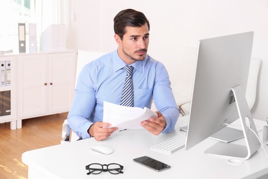 Photo of Handsome young man working with computer and papers at table in office