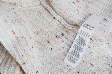 Photo of White clothing label with care information on beige warm sweater, top view. Space for text