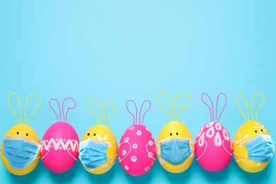 Image of COVID-19 pandemic. Colorful Easter eggs with cute bunny ears in protective masks and space for text on light blue background, flat lay