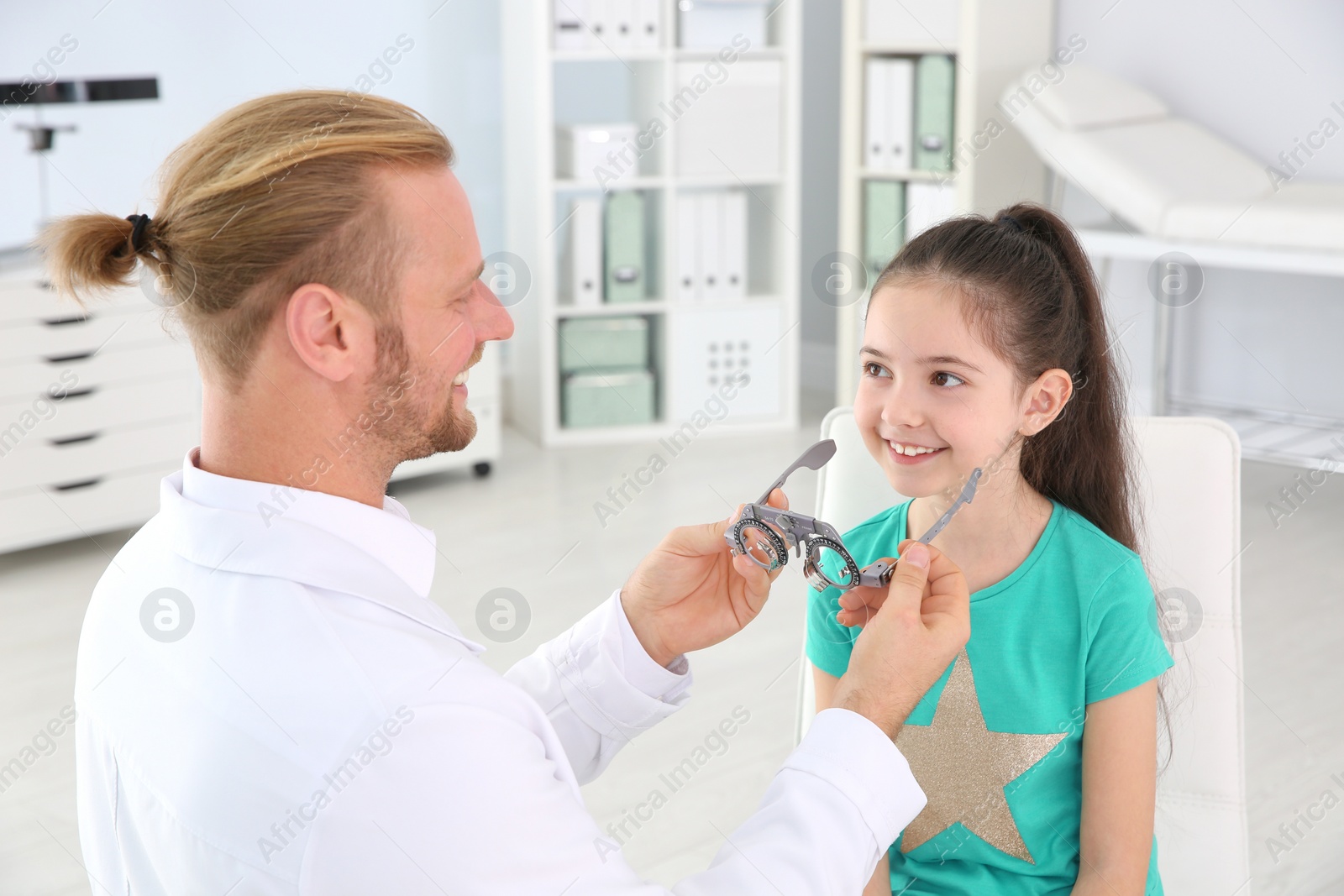 Photo of Children's doctor putting trial frame on little girl in clinic. Eye examination