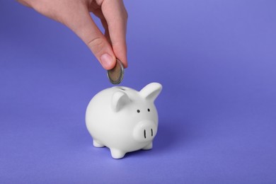 Woman putting coin into ceramic piggy bank on purple background, closeup with space for text. Financial savings