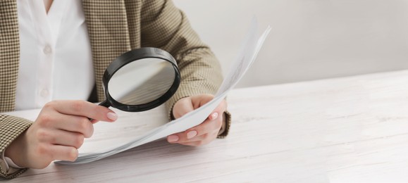 Image of Woman looking at documents through magnifier at white wooden table, closeup. Banner design with space for text