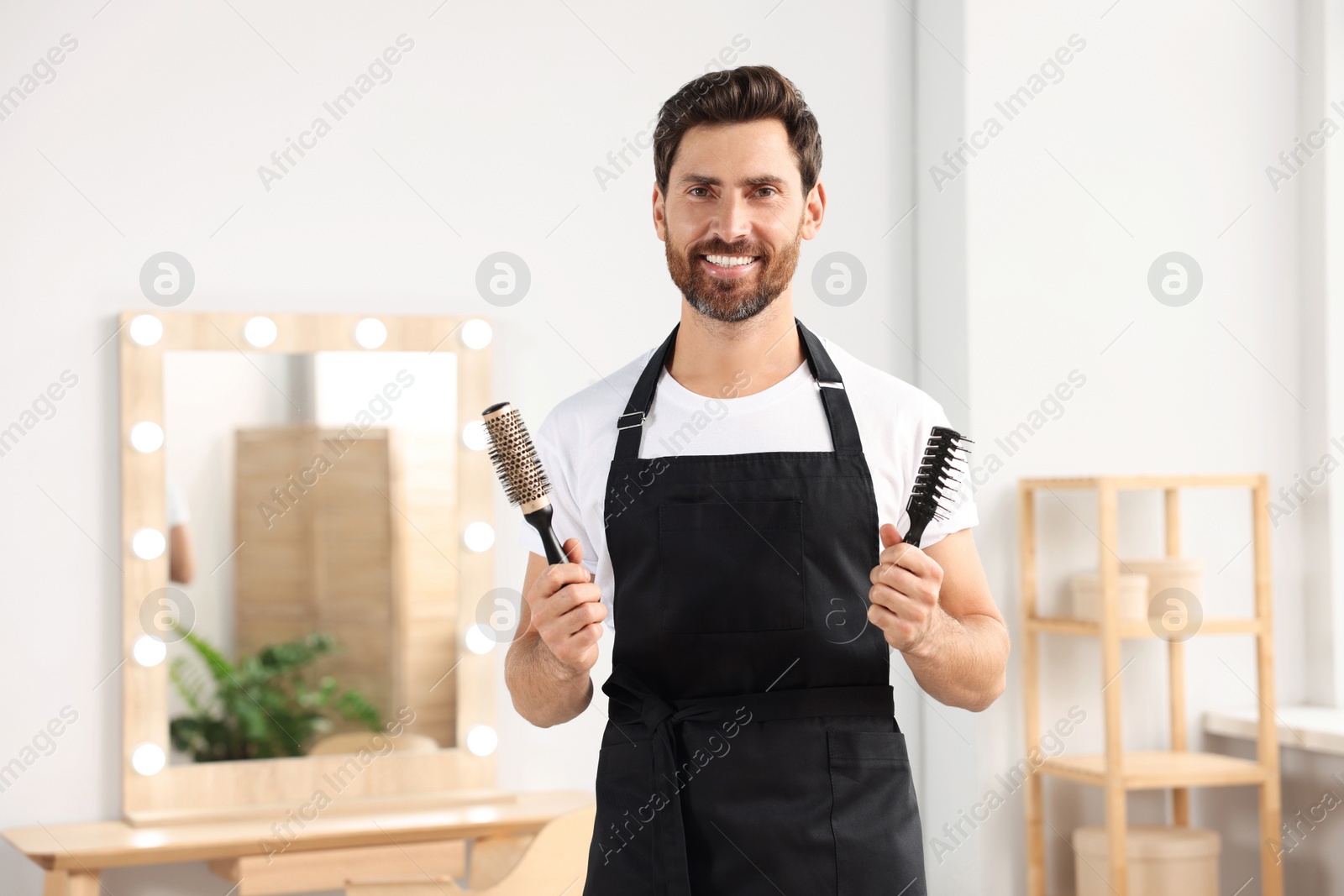 Photo of Smiling hairdresser in apron holding brushes near vanity mirror in salon, space for text