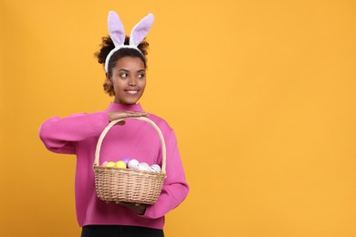 Photo of Happy African American woman in bunny ears headband holding wicker basket with Easter eggs on orange background, space for text