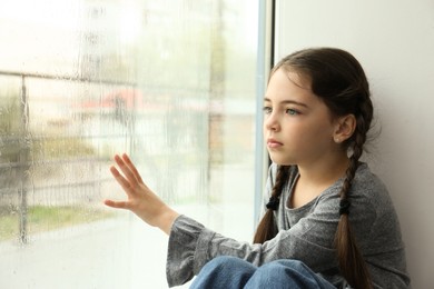 Photo of Sad little girl near window indoors on rainy day