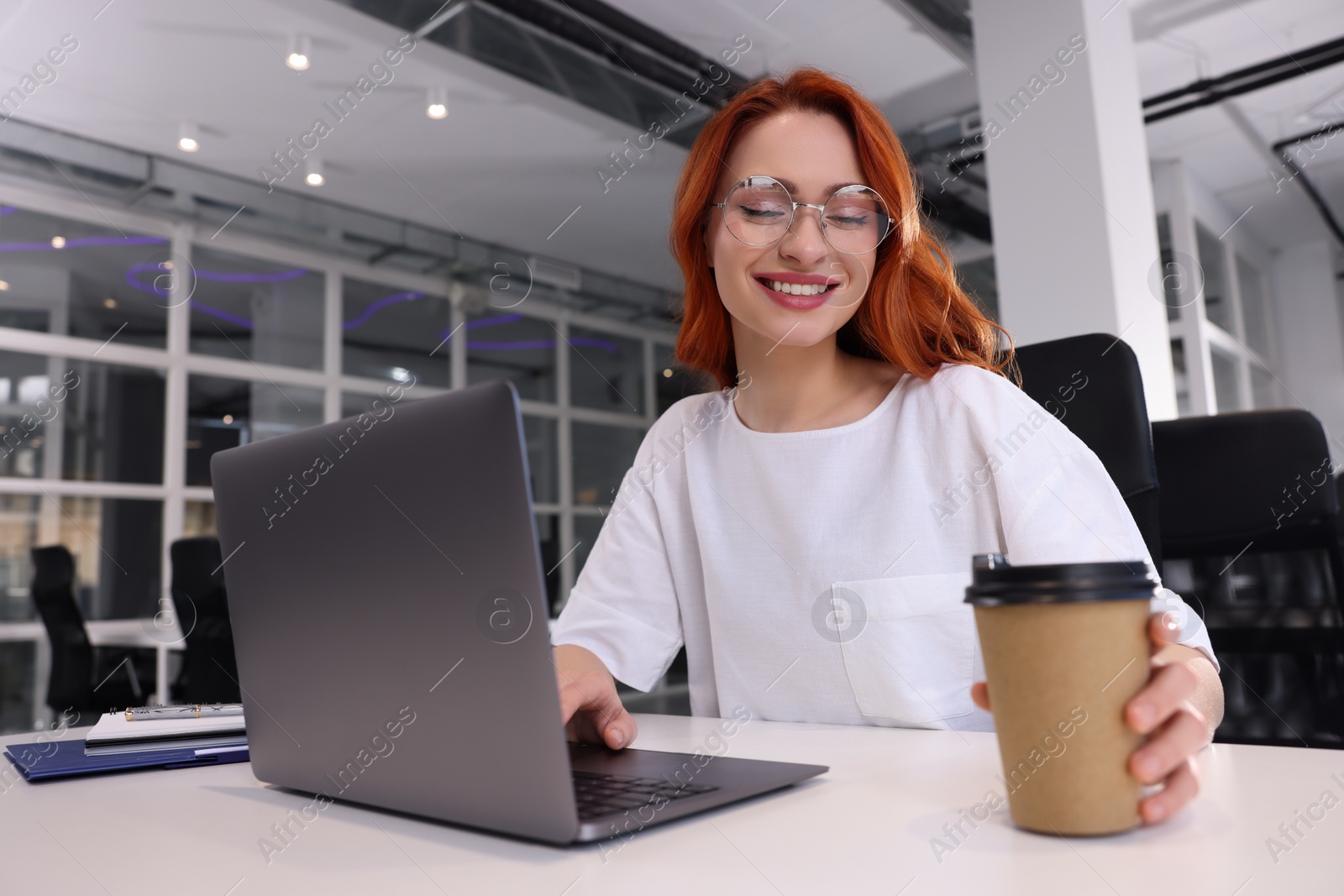 Photo of Happy woman with paper cup of coffee working on laptop at white desk in office
