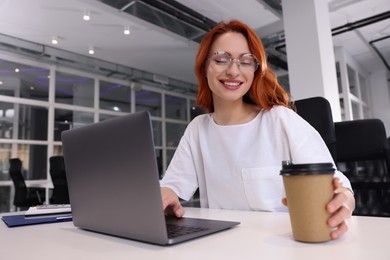 Photo of Happy woman with paper cup of coffee working on laptop at white desk in office