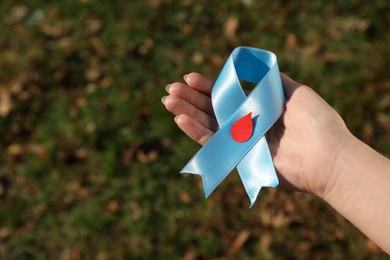 Photo of Woman holding light blue ribbon with paper blood drop outdoors, closeup. Diabetes awareness