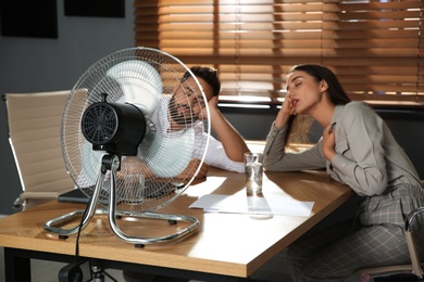 Photo of Office employees sitting in front of fan at workplace. Summer heat