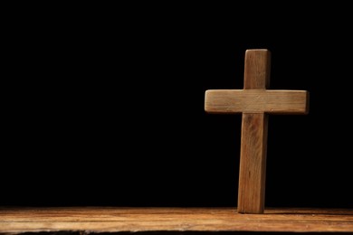 Cross on wooden table against black background, space for text. Religion of Christianity