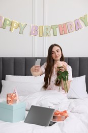 Photo of Beautiful young woman taking selfie with rose flowers, laptop and gift boxes on bed in room. Happy Birthday