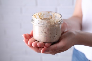 Photo of Woman holding glass jar with fresh sourdough starter on blurred background, closeup. Space for text