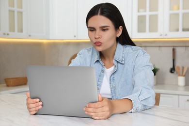 Young woman having video chat via laptop and sending air kiss at table in kitchen