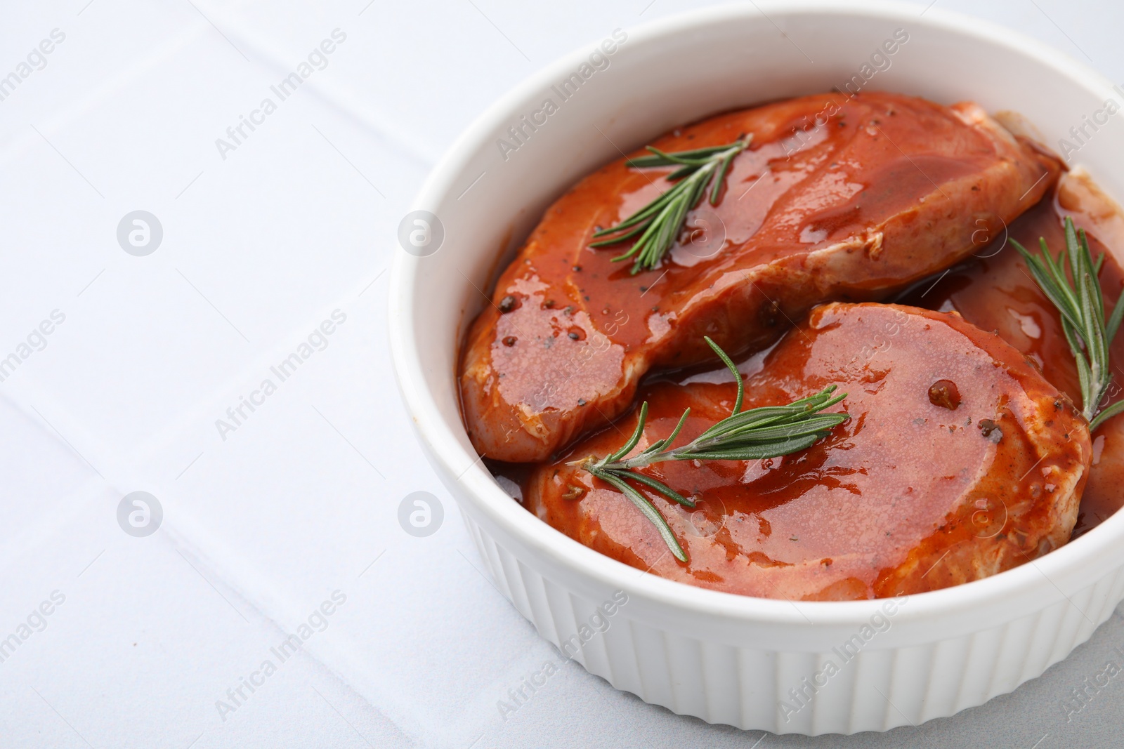 Photo of Raw marinated meat and rosemary in bowl on white tiled table, closeup. Space for text