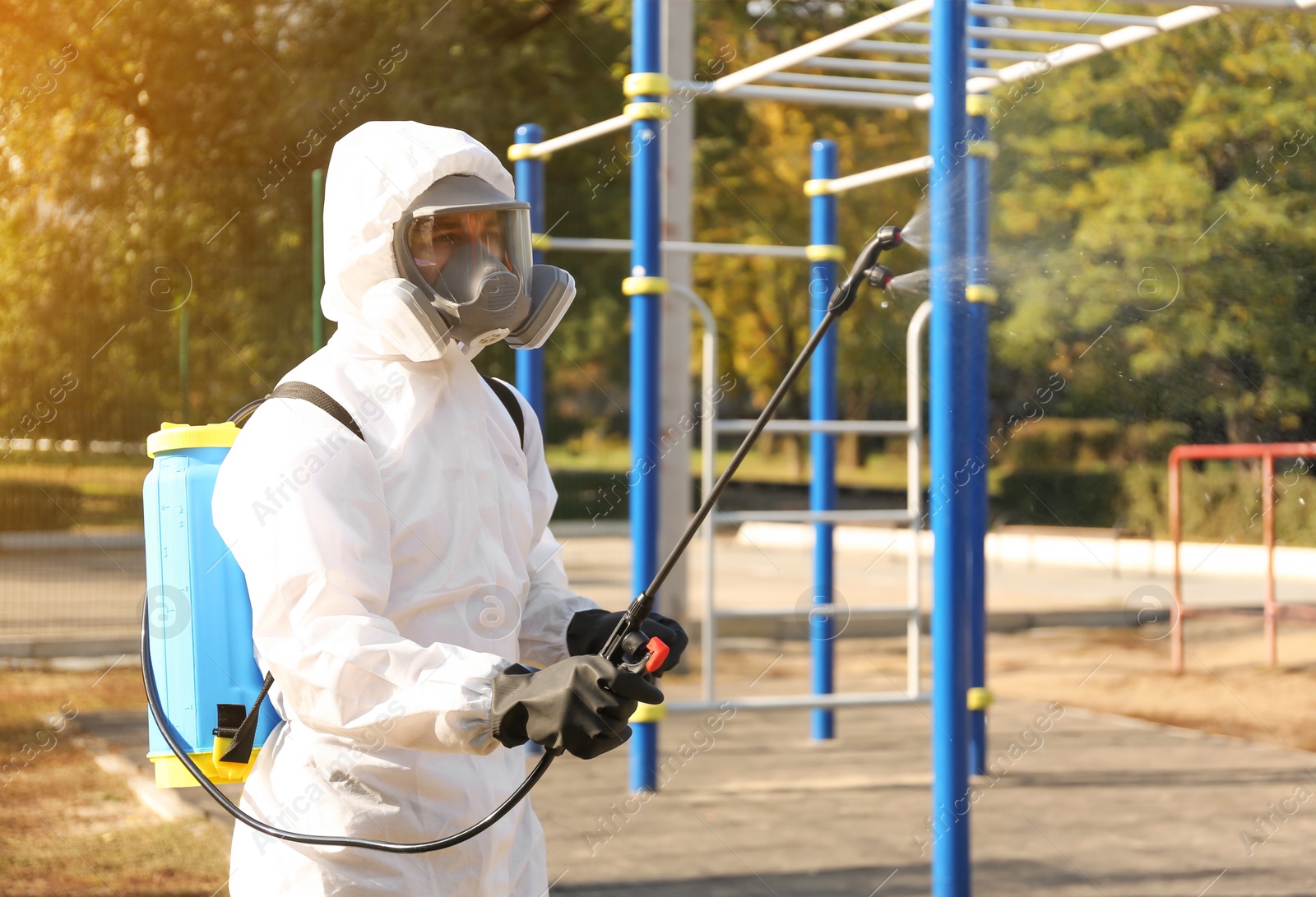 Photo of Man in hazmat suit spraying disinfectant around outdoor gym. Surface treatment during coronavirus pandemic