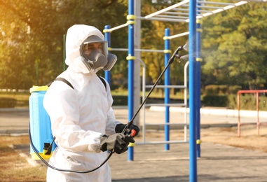 Photo of Man in hazmat suit spraying disinfectant around outdoor gym. Surface treatment during coronavirus pandemic