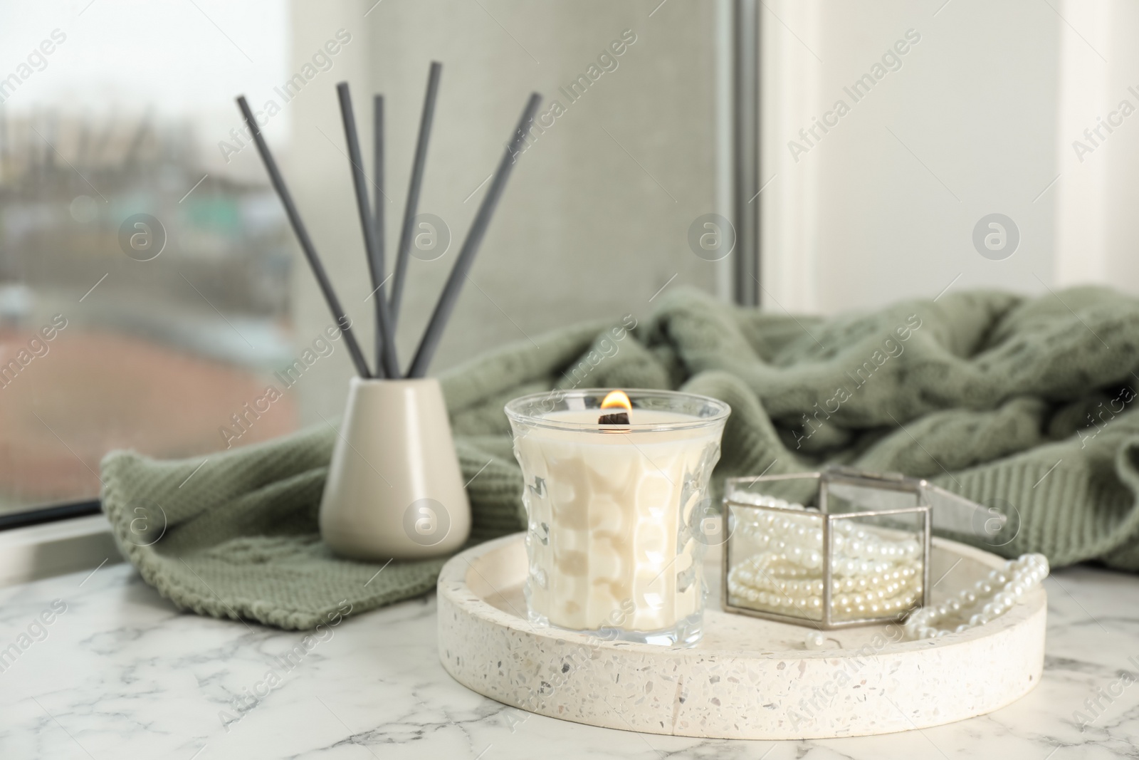 Photo of Tray with burning soy candle and beautiful bijouterie on white window sill indoors