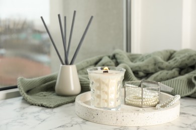 Tray with burning soy candle and beautiful bijouterie on white window sill indoors