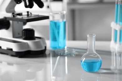 Different laboratory glassware with light blue liquid near microscope on table, space for text