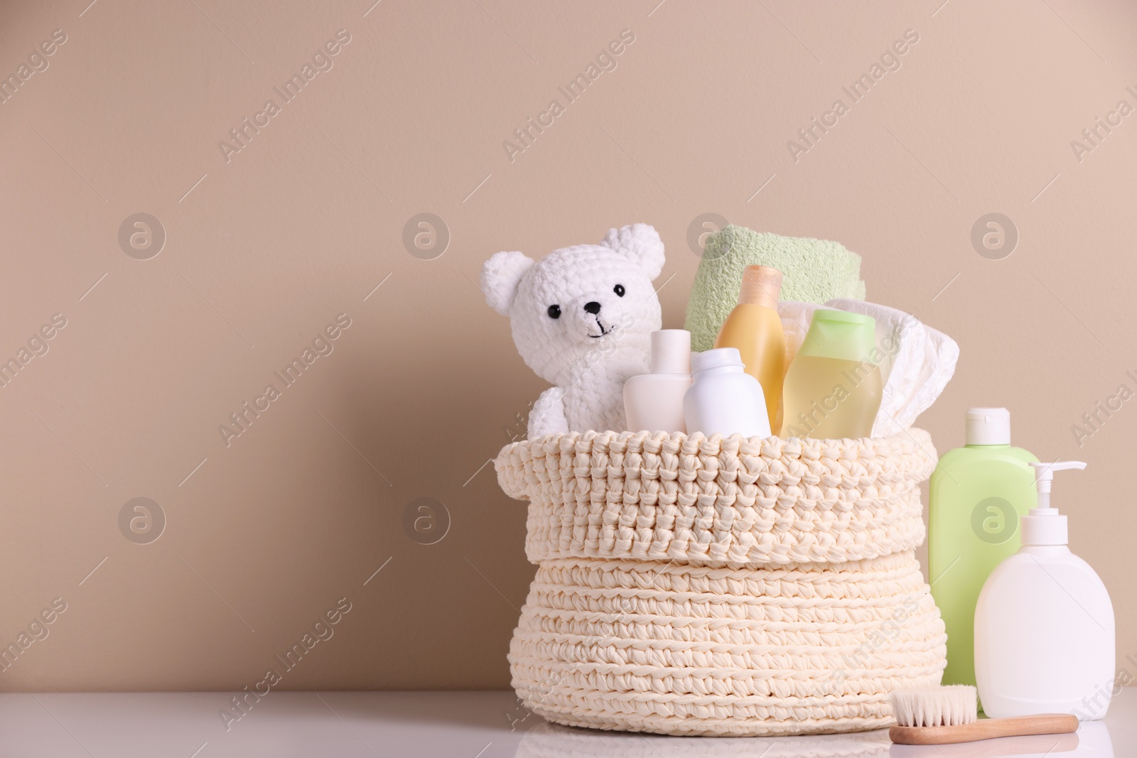 Photo of Knitted basket with baby cosmetic products, bath accessories and toy bear on white table against beige background. Space for text