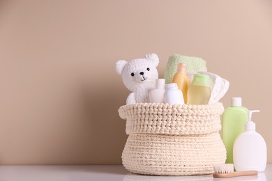 Photo of Knitted basket with baby cosmetic products, bath accessories and toy bear on white table against beige background. Space for text