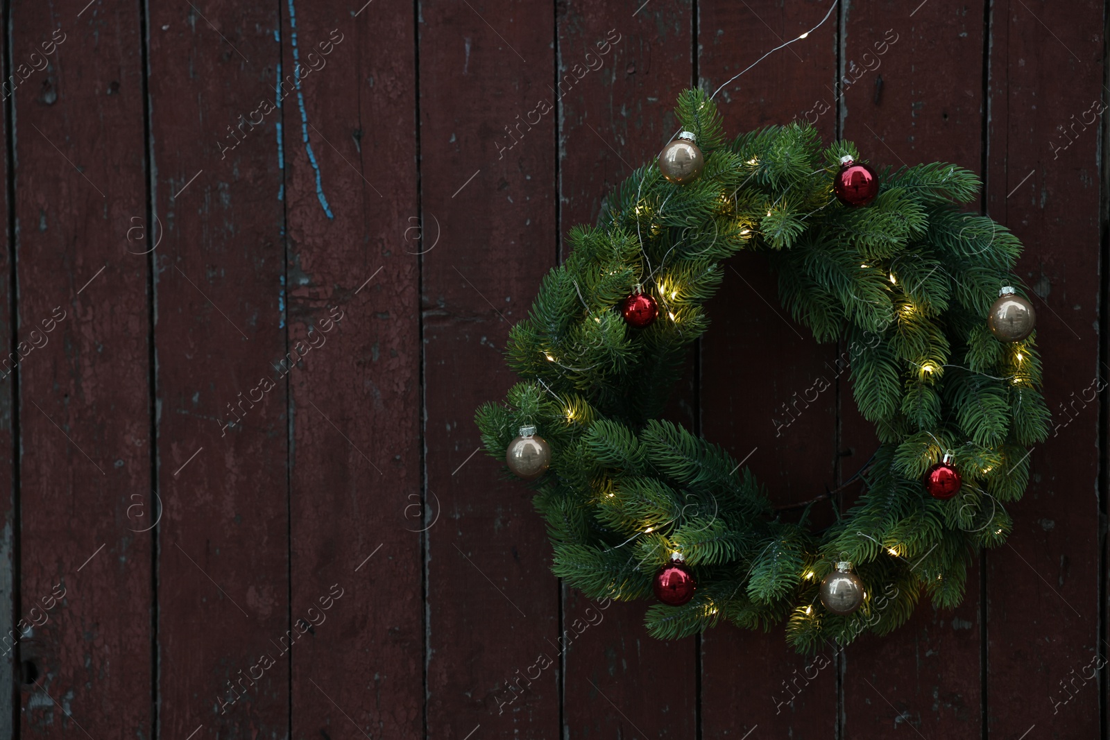 Photo of Beautiful Christmas wreath with baubles and string lights hanging on brown wooden wall, space for text