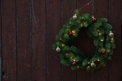 Photo of Beautiful Christmas wreath with baubles and string lights hanging on brown wooden wall, space for text