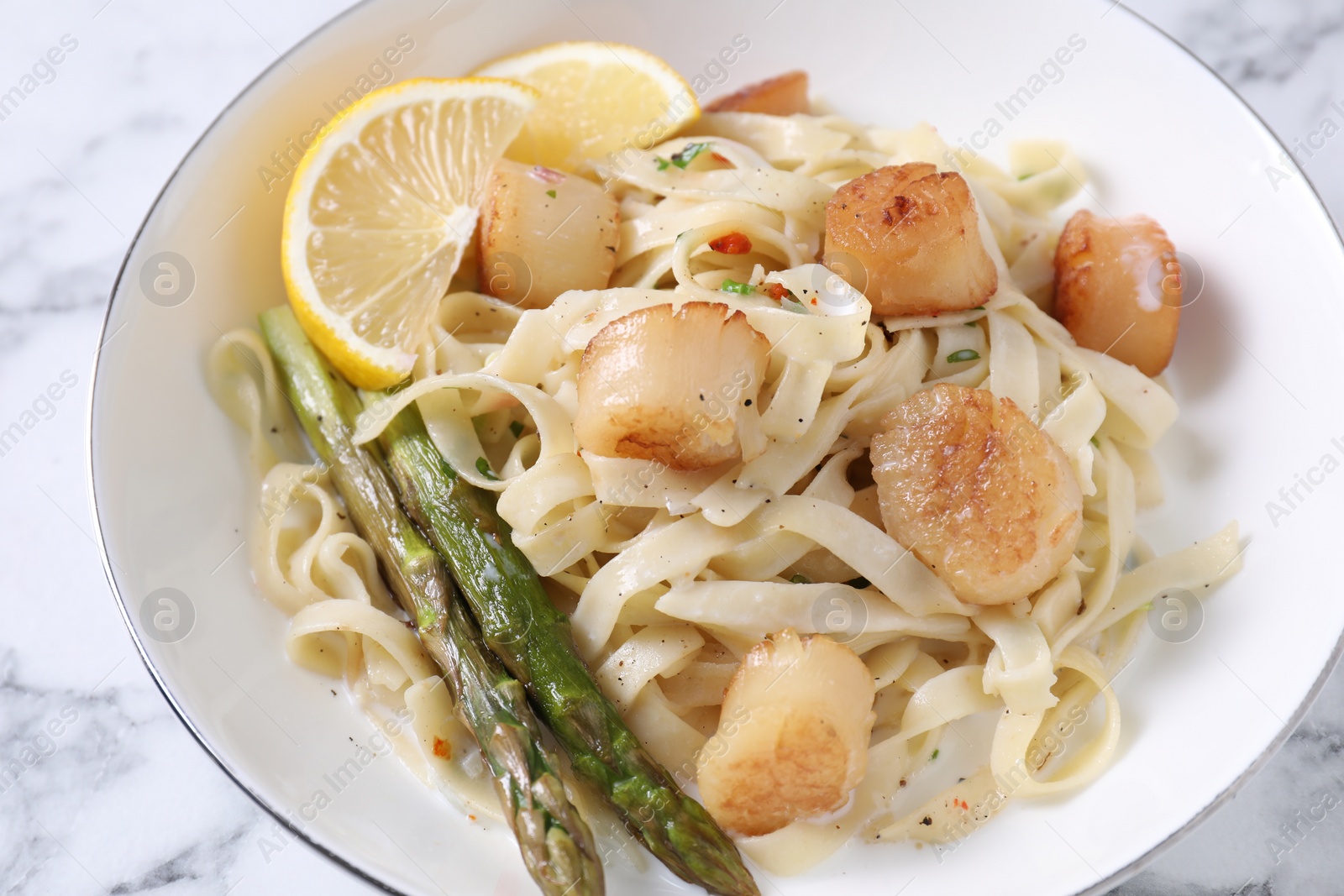 Photo of Delicious scallop pasta with asparagus and lemon on white marble table, closeup