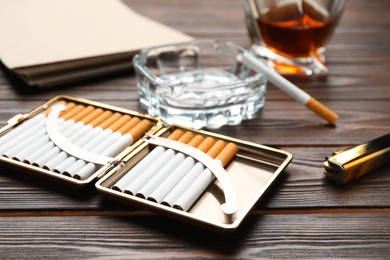Open case with tobacco filter cigarettes, ashtray, alcohol drink and lighter on wooden table, closeup