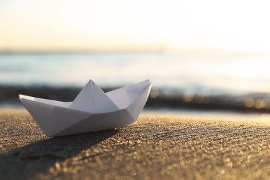 Photo of White paper boat on sand near sea at sunset, closeup. Space for text