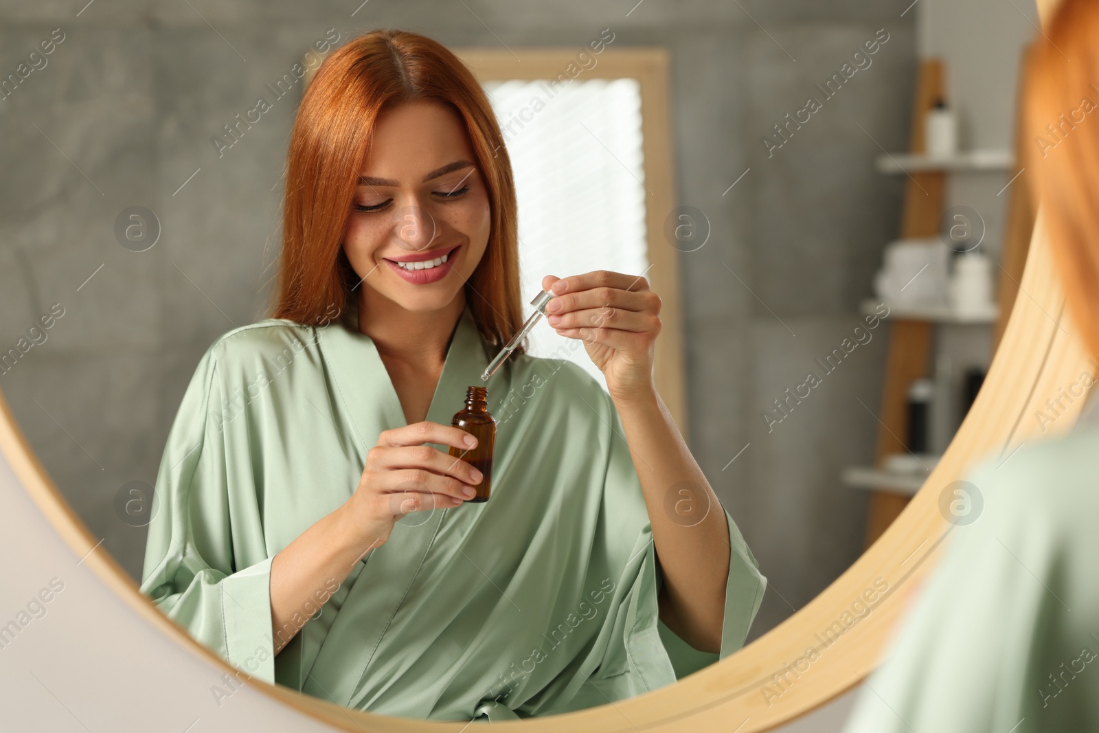 Photo of Beautiful young woman with bottle of cosmetic serum near mirror in bathroom