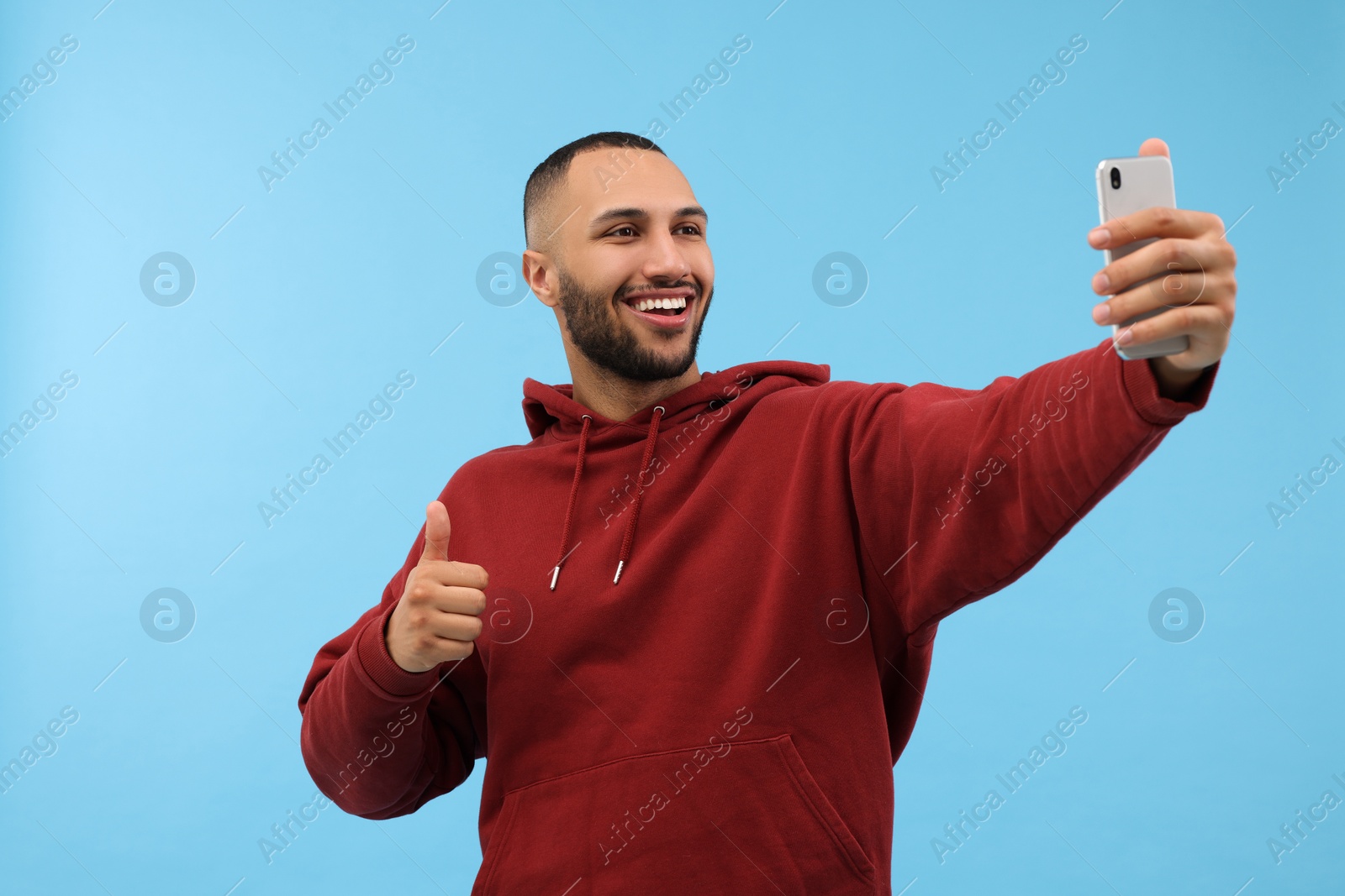 Photo of Smiling young man taking selfie with smartphone and showing thumbs up on light blue background