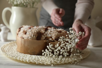 Photo of Woman decorating delicious Italian Easter dove cake (traditional Colomba di Pasqua) with flowers at white wooden table, closeup