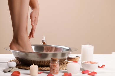 Photo of Woman soaking her foot in bowl with water and rose petals on white wooden surface, closeup. Pedicure procedure