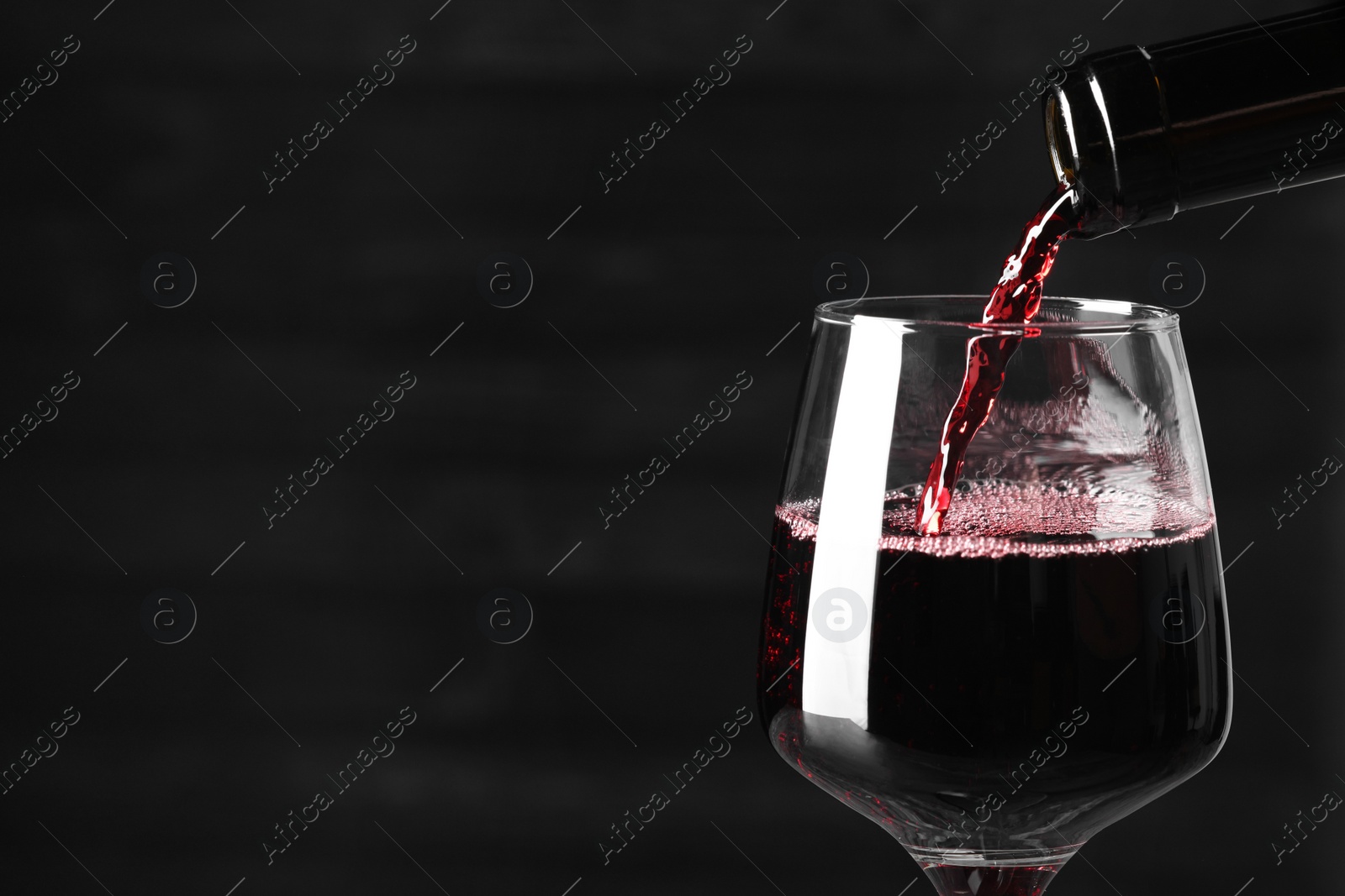 Photo of Pouring red wine into glass against dark background, closeup. Space for text