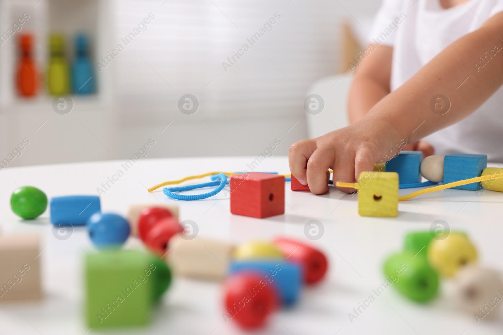 Photo of Motor skills development. Little boy playing with wooden pieces and string for threading activity at white table indoors, closeup