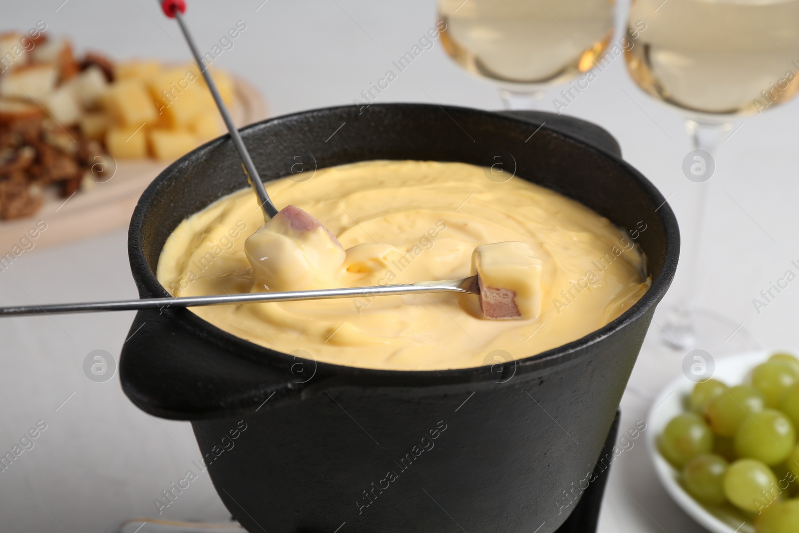 Photo of Dipping ham into fondue pot with tasty melted cheese at white table, closeup