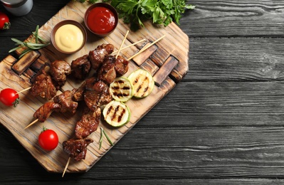 Photo of Board with barbecued meat, garnish and sauces on wooden background, top view. Space for text