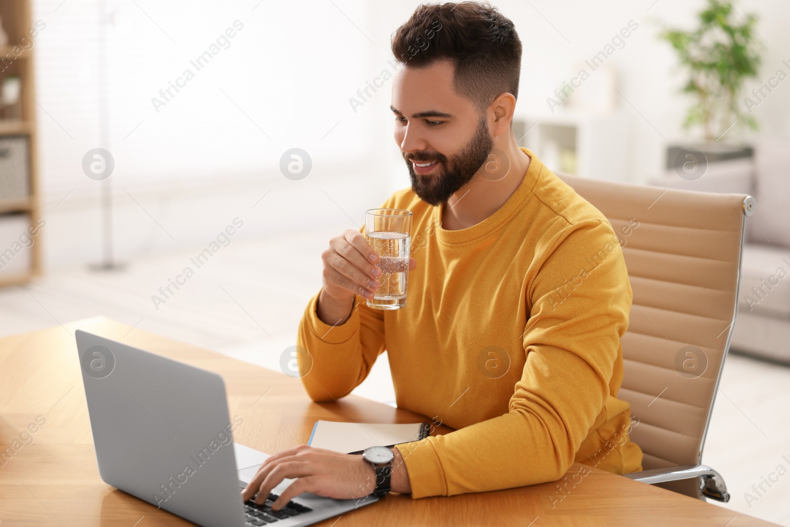 Photo of Young man with glass of water watching webinar at table in room