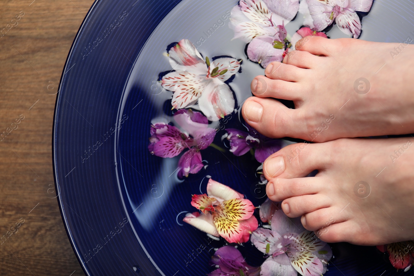 Photo of Woman putting her feet in bowl with water and flowers on wooden floor, top view. Spa treatment